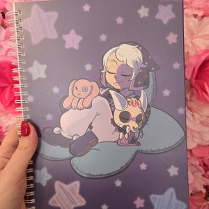 May include: A spiral-bound notebook with a purple cover featuring a cartoon illustration of a character sleeping on a cloud with a plush toy and a skull. The cover is decorated with stars and the text "Sweet Sakura Studio" and "calmeuniverse".