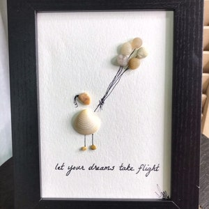 May include: A framed artwork featuring a seashell figure holding a bunch of balloons made from small stones. The text "let your dreams take flight" is written below the figure.