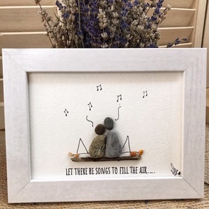 May include: A white framed artwork with a hand-drawn illustration of two figures sitting on a branch with musical notes around them. The text below reads "Let there be songs to fill the air..."
