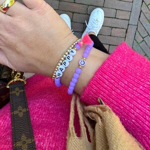 Könnte beinhalten: Nahaufnahme eines Handgelenks mit zwei Armbändern. Eines hat goldene Perlen und weiße Buchstabenperlen mit der Aufschrift "HAPPY". Das andere ist ein lila Perlenarmband mit einem Smiley-Anhänger und einer rosa Quaste.
