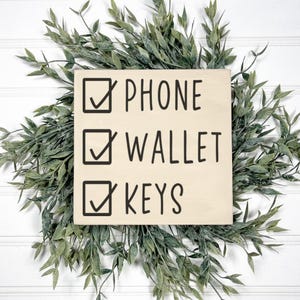 May include: A square wooden sign with the words "PHONE", "WALLET", and "KEYS" in black, each with a checkmark in a box. The sign is surrounded by a green leafy wreath, set against a white background.