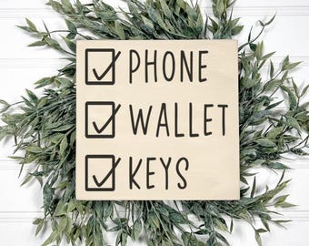 May include: A square wooden sign with the words "PHONE", "WALLET", and "KEYS" in black, each with a checkmark in a box. The sign is surrounded by a green leafy wreath, set against a white background.