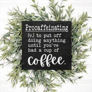 May include: A black wooden sign with white text that reads "Procaffeinating (v.) to put off doing anything until you've had a cup of coffee." The sign is surrounded by green leafy sprigs, set against a white background.