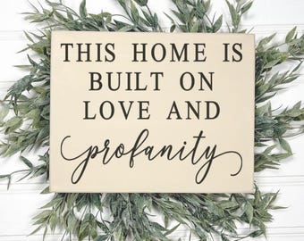 May include: A wooden sign with the text "This home is built on love and profanity" in black lettering on a cream background.
