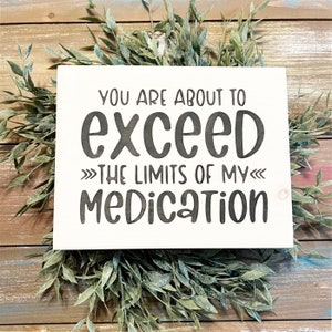 May include: A white wooden sign with the phrase "YOU ARE ABOUT TO EXCEED THE LIMITS OF MY MEDICATION" in black lettering. The sign is surrounded by green leafy sprigs, set against a wooden background.