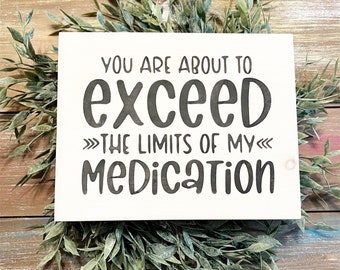 May include: A white wooden sign with the phrase "YOU ARE ABOUT TO EXCEED THE LIMITS OF MY MEDICATION" in black lettering. The sign is surrounded by green leafy sprigs, set against a wooden background.
