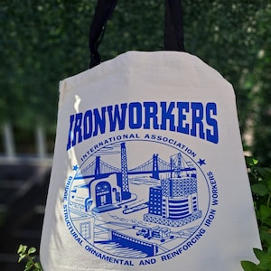 May include: A white canvas tote bag with blue text and a graphic of a bridge and buildings. The text reads "Ironworkers International Association Bridge, Structural, Ornamental and Reinforcing Iron Workers".