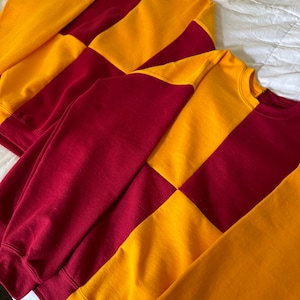 Pode incluir: Duas sweatshirts com um padrão xadrez em vinho e dourado. As sweatshirts estão dobradas uma em cima da outra.