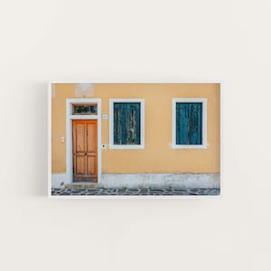 Puede incluir: Un edificio amarillo con una puerta de madera y dos ventanas con contraventanas azules. El edificio tiene un marco blanco alrededor de las ventanas y la puerta. El edificio está frente a una pared blanca.