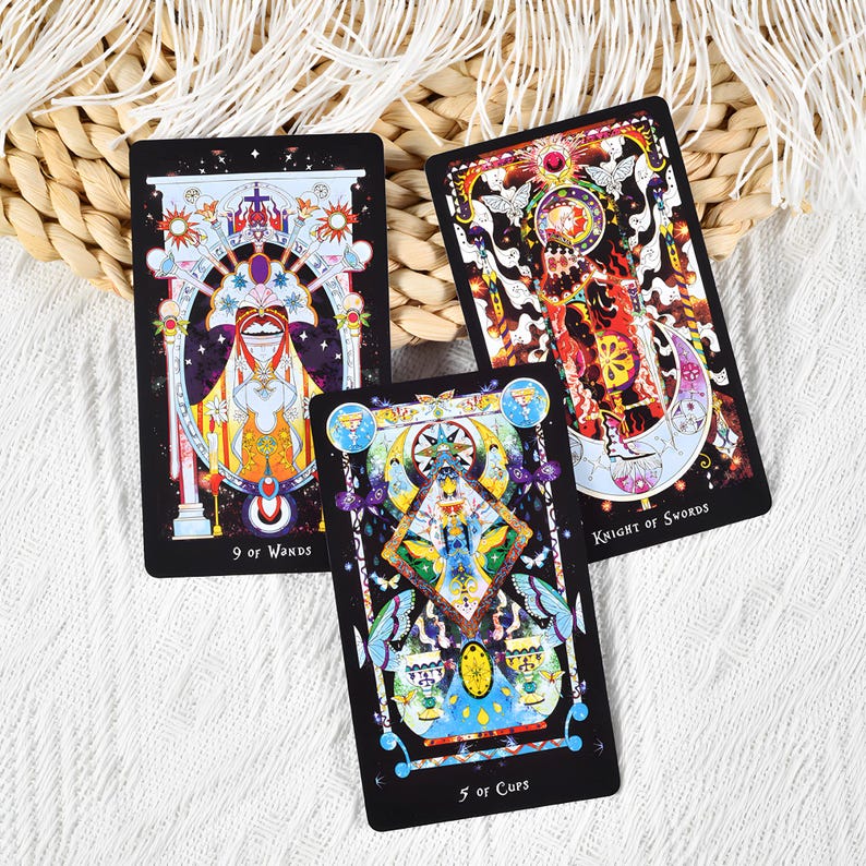 May include: Three tarot cards with colorful illustrations on a white surface. The cards are black with detailed artwork featuring celestial and symbolic imagery. The cards are labeled "9 of Wands", "Knight of Swords", and "5 of Cups".