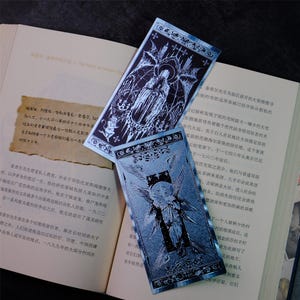 May include: Two ornate, blue-toned bookmarks with detailed illustrations of figures with wings and halos. The bookmarks are resting on an open book with Chinese text. The bookmarks have a gothic or fantasy theme.