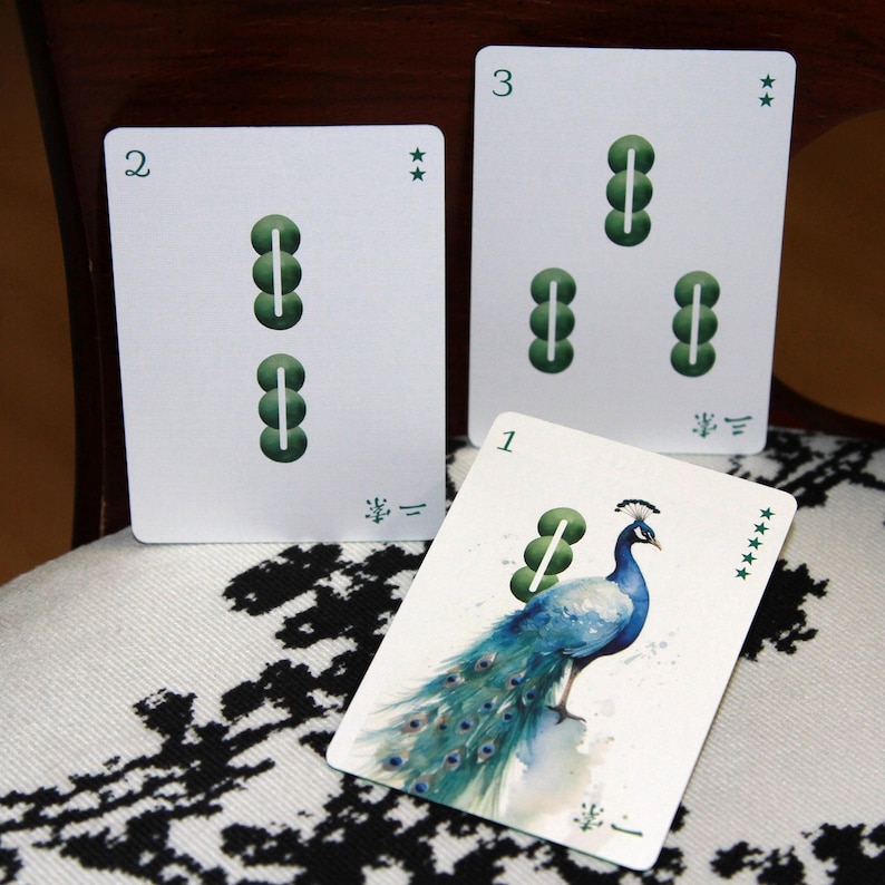 Pu&ograve; includere: Tre carte da gioco con sfondi bianchi. Le carte presentano simboli e numeri verdi. Una carta ha un'illustrazione di pavone ad acquerello. Le carte sono disposte su una superficie a motivi bianchi e neri.