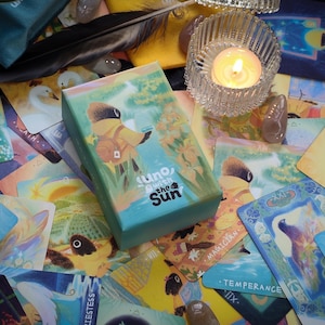 May include: A teal box of "Uno with the Sun" tarot cards rests on a collection of colorful tarot cards. A lit candle in a glass holder and a black feather are also visible. The box features an illustration of a figure in a yellow coat.