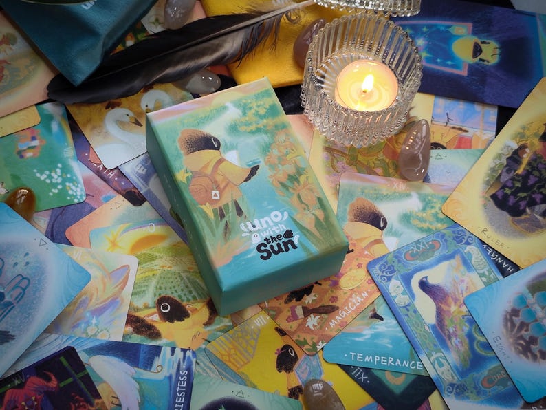 May include: A teal box of "Uno with the Sun" tarot cards rests on a collection of colorful tarot cards. A lit candle in a glass holder and a black feather are also visible. The box features an illustration of a figure in a yellow coat.