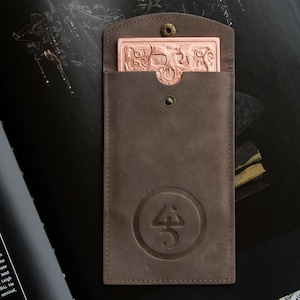 May include: A brown leather sleeve with a snap closure and a circular embossed logo. Inside the sleeve is a rectangular copper-colored metal card featuring an intricate relief design of a skull and swirling patterns on its surface.