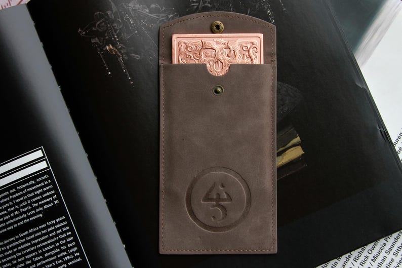 May include: A brown leather sleeve with a snap closure and a circular embossed logo. Inside the sleeve is a rectangular copper-colored metal card featuring an intricate relief design of a skull and swirling patterns on its surface.