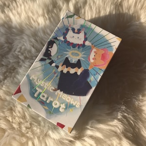 K&ouml;nnte beinhalten: Eine Tarotkartenbox mit dem Text "Mystic Meows Tarot" in Wei&szlig;. Die Box zeigt Illustrationen von Katzenfiguren mit Heiligenscheinen und farbenfrohen Akzenten. Der Hintergrund ist hellblau mit radialen Linien.