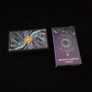 May include: A deck of Broken Mirror Tarot cards featuring a rectangular box with a purple cover, a crescent moon, and the Roman numeral V. Beside it lies a card displaying a gold starburst design surrounded by swirling blue and black patterns.