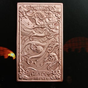 May include: A rectangular copper-toned metal card featuring an intricate embossed design of a cosmic scene with tentacles, celestial bodies, and a planet. The bottom section displays the text THE FOOL in raised capital letters within a decorative border.