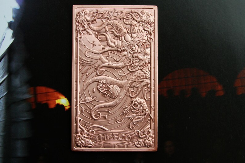 May include: A rectangular copper-toned metal card featuring an intricate embossed design of a cosmic scene with tentacles, celestial bodies, and a planet. The bottom section displays the text THE FOOL in raised capital letters within a decorative border.