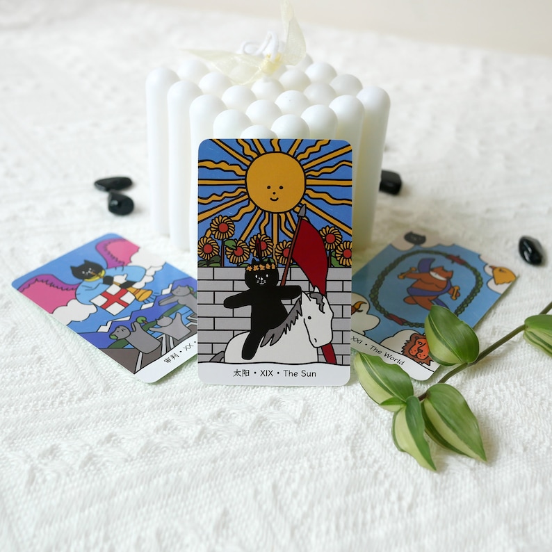 Puede incluir: Una carta del tarot con un gato negro montando un caballo blanco, con una bandera roja y un sol amarillo con una cara sonriente. La carta se titula "El Sol" y tiene el n&uacute;mero XIX.