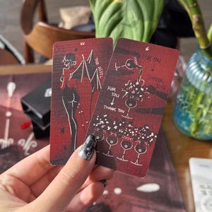 Puede incluir: Dos cartas de tarot con fondo rojo. Una carta presenta una figura de diablo, la otra tiene copas de vino y el texto "FOR YOU" y "THANK YOU". Las cartas se sostienen en una mano.