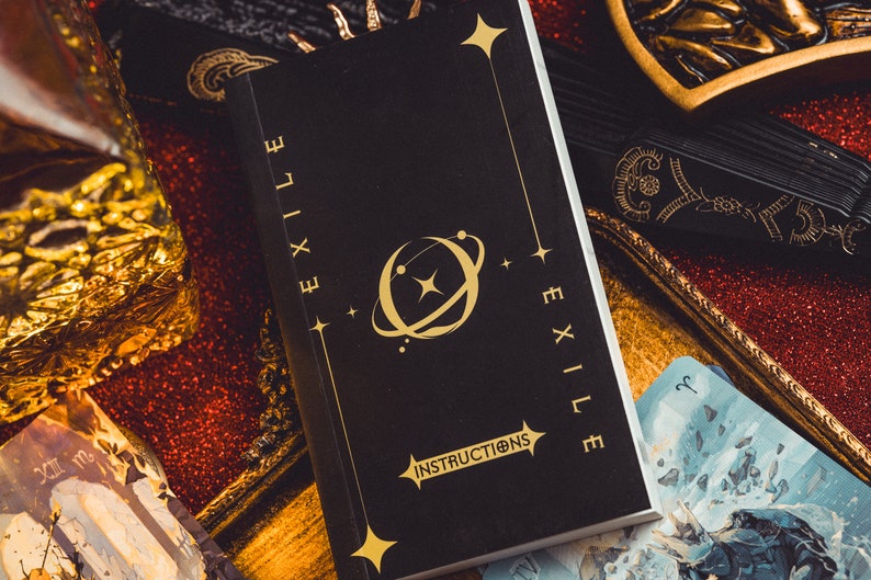 Op de afbeelding: Een zwart boek met de gouden tekst "EXILE" op de rug en de kaft. De kaft toont een gouden hemels ontwerp en het woord "INSTRUCTIONS". Het boek is omringd door andere tarotkaarten en decoratieve items.