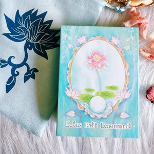 Może przedstawiać: Talia kart zatytułowana "Lotus Path Lenormands" z akwarelową ilustracją różowego kwiatu lotosu w owalnej ramie. Tło jest jasnoniebieskie z kwiatowymi akcentami. Jasnoniebieska tkanina z ciemnoniebieskim wzorem kwiatu lotosu w tle.