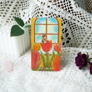 May include: A tarot card with a yellow, orange, and red tulip design. A woman in a white dress sits in a red tulip. The card is titled "The Sun" and has a star symbol.