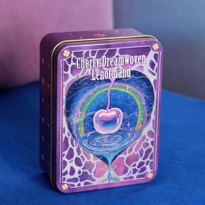 May include: A rectangular purple metal tin featuring the text Cherry Dreamwoven Lenormand. The front design displays a glowing cherry suspended above a swirling vortex, framed by a rainbow and a purple and white abstract pattern on the edges.