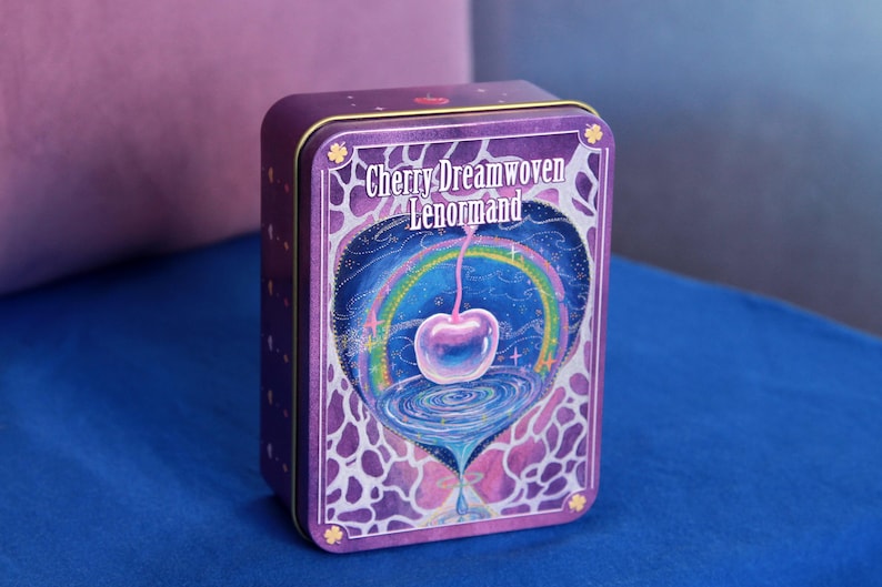 May include: A rectangular purple metal tin featuring the text Cherry Dreamwoven Lenormand. The front design displays a glowing cherry suspended above a swirling vortex, framed by a rainbow and a purple and white abstract pattern on the edges.