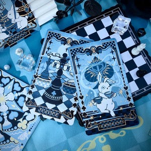 Puede incluir: Cartas de tarot con un diseño caprichoso, que presentan un conejo blanco y elementos de ajedrez en tonos azules y negros. Las cartas muestran las palabras "Justice" y "Temperance". También se ve un pequeño tablero de ajedrez y elementos decorativos.
