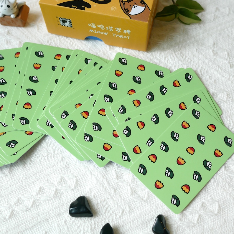 Puede incluir: Una baraja de cartas de tarot verde con un patr&oacute;n de flores blancas y huevos naranjas. La caja de las cartas est&aacute; en el fondo y dice "喵喵塔羅 MIAOW TAROT" en chino e ingl&eacute;s.