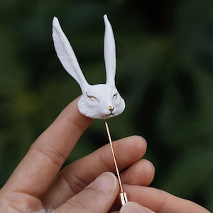 May include: A white rabbit head lapel pin with gold detailing on the eyes and nose. The pin features a long, thin, gold-coloured needle. The rabbit's ears are long and upright. The pin is set against a blurred green backdrop.