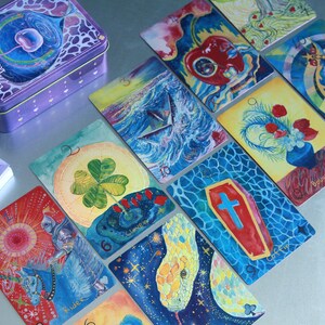 May include: A deck of Cherry Dreamwoven Lenormand cards featuring colourful, detailed illustrations on each card, including a clover, a coffin, a snake, and a knight. The cards are displayed alongside a purple metal storage tin with matching artwork.