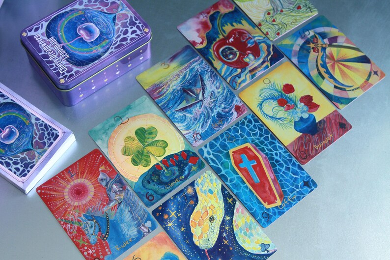 May include: A deck of Cherry Dreamwoven Lenormand cards featuring colourful, detailed illustrations on each card, including a clover, a coffin, a snake, and a knight. The cards are displayed alongside a purple metal storage tin with matching artwork.