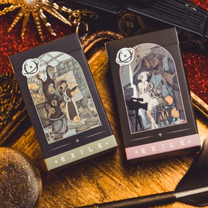 Op de afbeelding: Twee zwarte EXILE+ kaartenboxen met ge&iuml;llustreerde sc&egrave;nes, een met een heksenachtige figuur en de andere met een filmset. De dozen hebben een wit planeetlogo en liggen op een gouden dienblad met een rode glitterachtergrond.