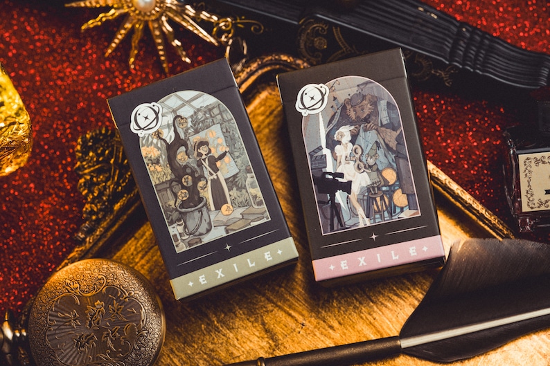 Op de afbeelding: Twee zwarte EXILE+ kaartenboxen met ge&iuml;llustreerde sc&egrave;nes, een met een heksenachtige figuur en de andere met een filmset. De dozen hebben een wit planeetlogo en liggen op een gouden dienblad met een rode glitterachtergrond.