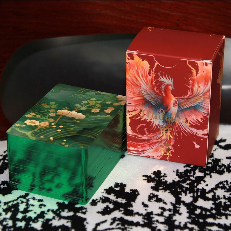 Pu&ograve; includere: Due scatole decorative. Una &egrave; verde scuro con un motivo floreale, l'altra &egrave; rossa con un'illustrazione di una fenice. La fenice ha piume arancioni, gialle e blu. Le scatole sono su una superficie a motivi bianchi e neri.