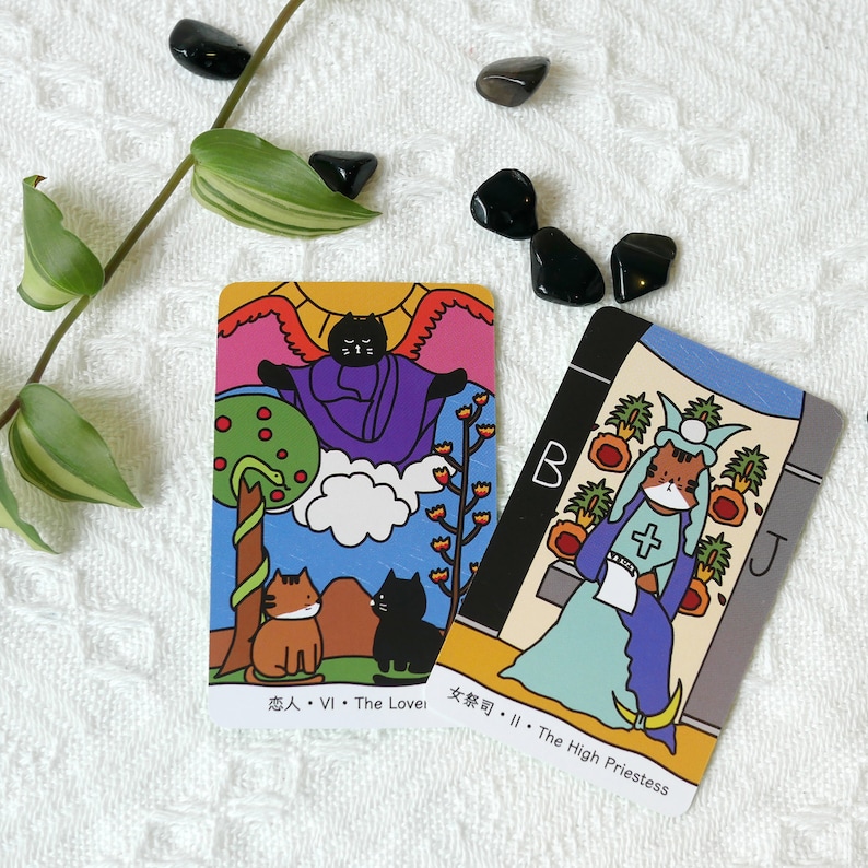 Puede incluir: Dos cartas de tarot con ilustraciones coloridas de gatos. La carta de la izquierda es la carta de Los Enamorados, y la carta de la derecha es la carta de La Sacerdotisa.