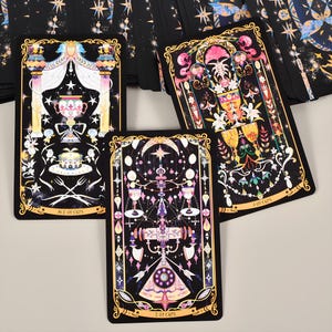 May include: Three tarot cards with ornate designs are displayed. The cards feature black backgrounds with gold borders and colorful illustrations of chalices, stars, and floral elements. The text "ACE OF CUPS" and "2 OF CUPS" and "3 OF CUPS" are visible.
