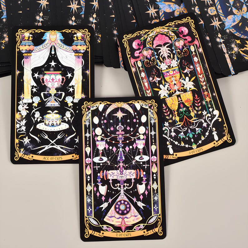 May include: Three tarot cards with ornate designs are displayed. The cards feature black backgrounds with gold borders and colorful illustrations of chalices, stars, and floral elements. The text "ACE OF CUPS" and "2 OF CUPS" and "3 OF CUPS" are visible.
