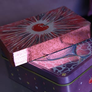 May include: A deck of playing cards with metallic pink edges resting on a rectangular purple tin box. The top card features a red cherry illustration with radiating white lines. The purple tin displays a cherry graphic and small star accents.