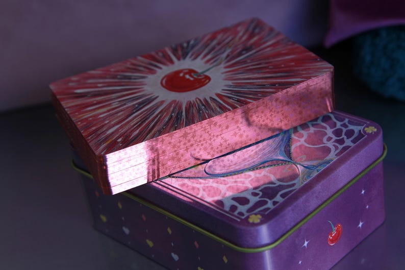 May include: A deck of playing cards with metallic pink edges resting on a rectangular purple tin box. The top card features a red cherry illustration with radiating white lines. The purple tin displays a cherry graphic and small star accents.
