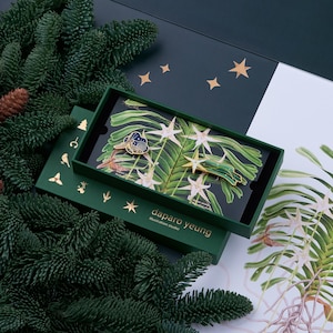 May include: A green box with a gold border and the text "daparo yeung illustration studio" on the front. The box contains two enamel pins, one with a blue butterfly and the other with a gold star. The pins are on a background of green leaves and white flowers.