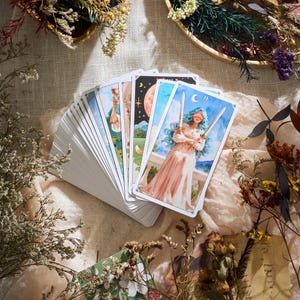May include: A deck of tarot cards with colorful illustrations, fanned out on a beige cloth. The cards feature images of figures, celestial bodies, and landscapes. Dried flowers and decorative elements surround the deck, creating a mystical aesthetic.