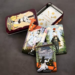May include: A tarot card set featuring illustrated cards and a burgundy metal tin. The tin has an illustration of a person with red hair. Several cards are visible, including the Justice and 8 of Pentacles cards.