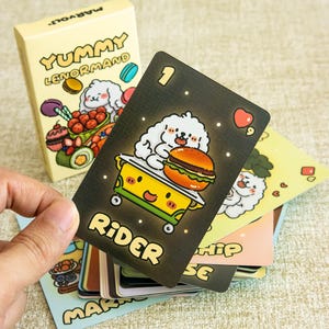 May include: A deck of Yummy Lenormand cards featuring a cream-colored box with cartoon food and a white dog. The top card is black with the number 1, a white dog on a yellow cart with a burger, and the text Rider. Other cards show colorful illustrations.