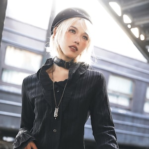 May include: A person wearing a black beret, choker, and long-sleeved shirt. The shirt has a ruffled collar and cuffs. A pendant necklace hangs from the neck. The background includes a train car and overhead beams.