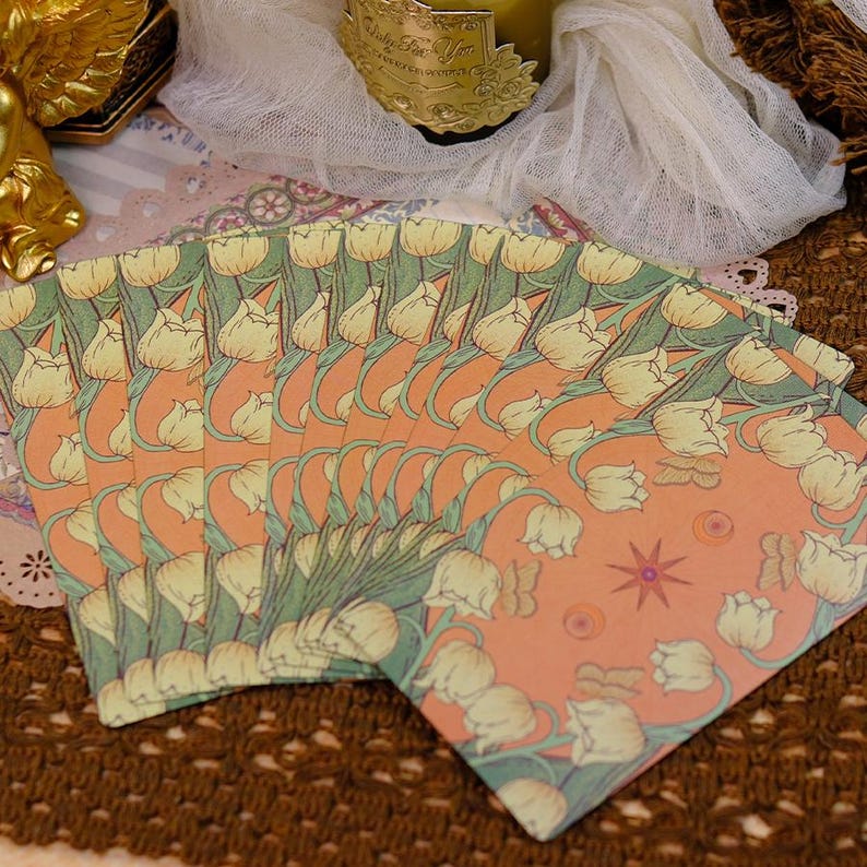K&ouml;nnte beinhalten: Ein Stapel Briefpapier mit floralem Design. Die Karten zeigen ein sich wiederholendes Muster aus cremefarbenen Tulpen mit gr&uuml;nen Bl&auml;ttern auf einem pfirsichfarbenen Hintergrund. Das Briefpapier liegt auf einer braunen, strukturierten Oberfl&auml;che.